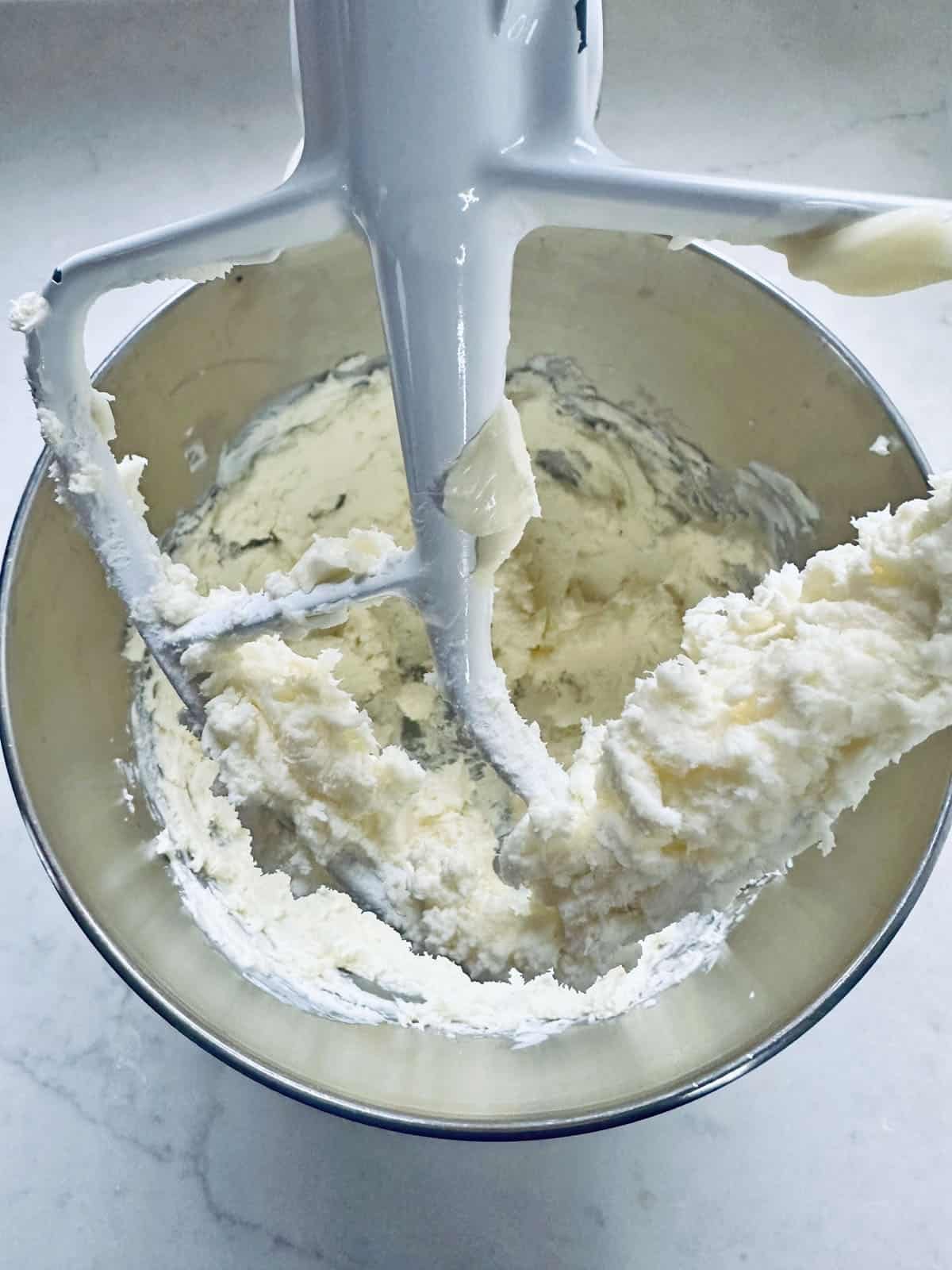 A close-up of a stand mixer with a paddle attachment mixing a creamy, pale batter in a metal bowl on a white countertop.