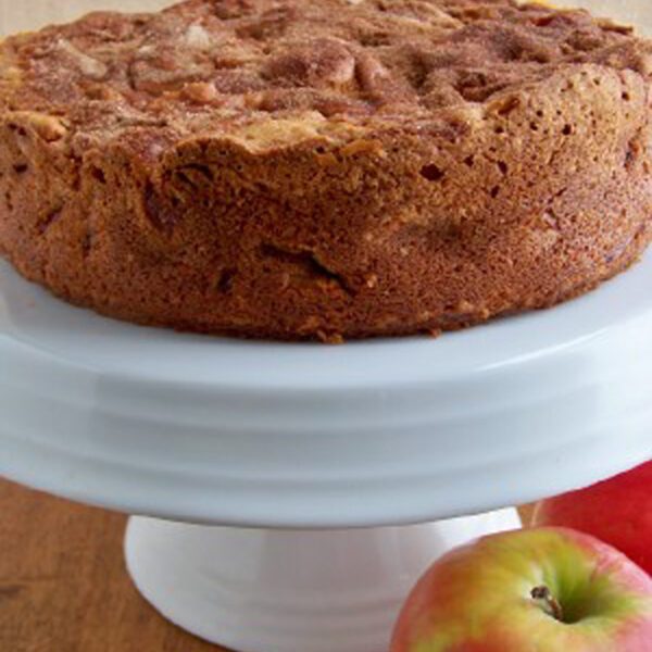 A round apple cake with a golden-brown crust sits on a white cake stand, with two fresh apples placed beside it on a wooden surface.