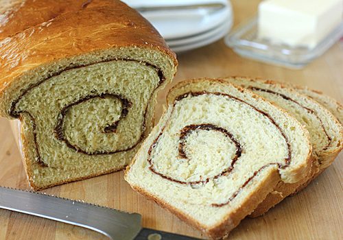 A loaf of cinnamon swirl bread with two slices cut, showing the spiral pattern inside. A knife, plates, and a dish of butter are in the background on a wooden surface.