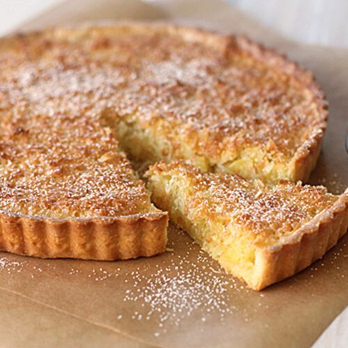 A golden-brown tart with a slice cut out sits on brown parchment paper. The tart is dusted with powdered sugar, and a metal sieve with powdered sugar is nearby.
