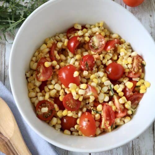 A white bowl filled with corn, cherry tomato halves, and herbs sits on a wooden table next to a wooden spoon, sprigs of fresh herbs, and loose cherry tomatoes.