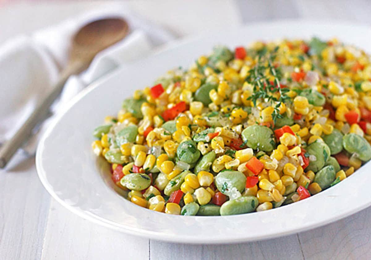 A white plate filled with succotash made of corn, lima beans, and diced red bell peppers, garnished with herbs. A wooden spoon and napkin are in the blurred background.
