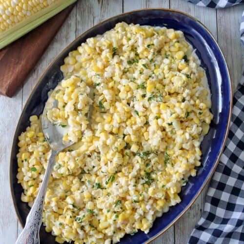 A blue oval platter filled with Corn Salad with Buttermilk and Herbs, topped with chopped herbs, with a serving spoon resting on the dish. Fresh corn on the cob and a checkered cloth sit nearby on a wooden surface.