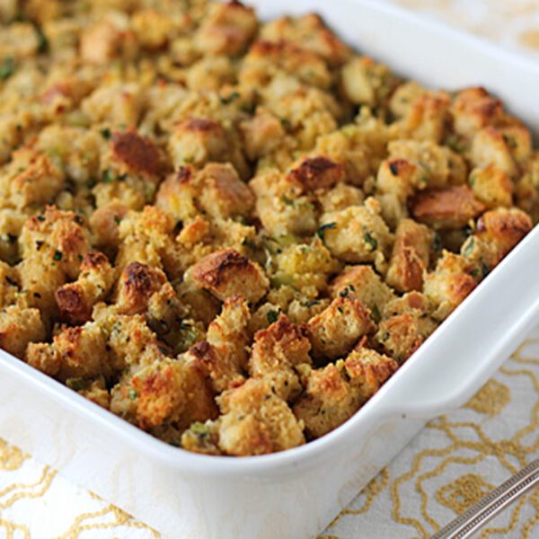 A white baking dish filled with golden-brown, baked stuffing made of cubed bread and herbs sits on a patterned tablecloth next to a metal serving spoon.