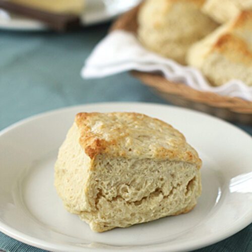 A golden, flaky biscuit sits on a white plate, placed on a light blue cloth. In the background, a basket of more biscuits and a dish of butter are visible.