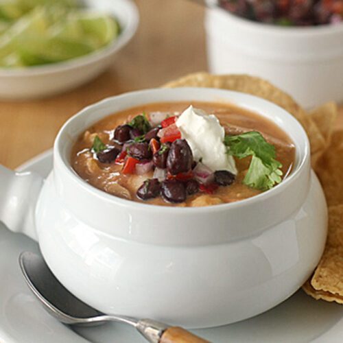 A bowl of creamy soup topped with black beans, diced tomatoes, cilantro, and a dollop of sour cream, served with tortilla chips on the side. A spoon rests on the plate, with lime wedges and salsa in the background.