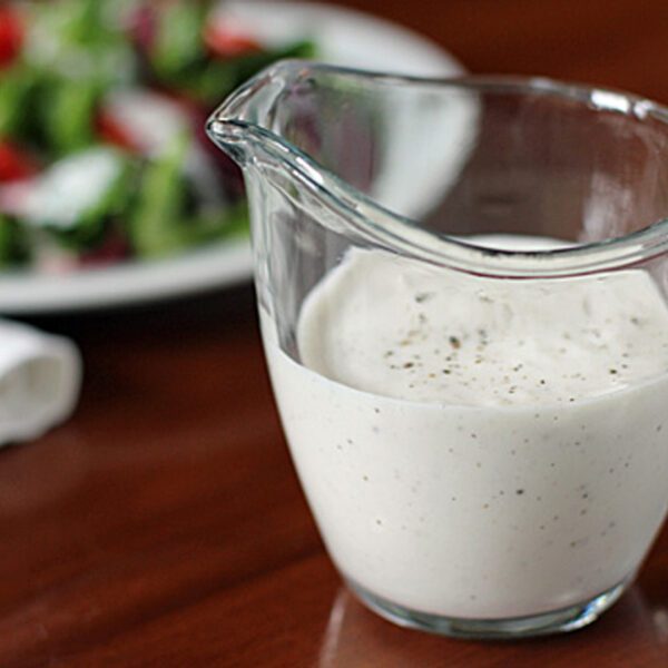 A glass pitcher filled with creamy white salad dressing sits on a wooden table, with a blurred plate of fresh salad in the background.