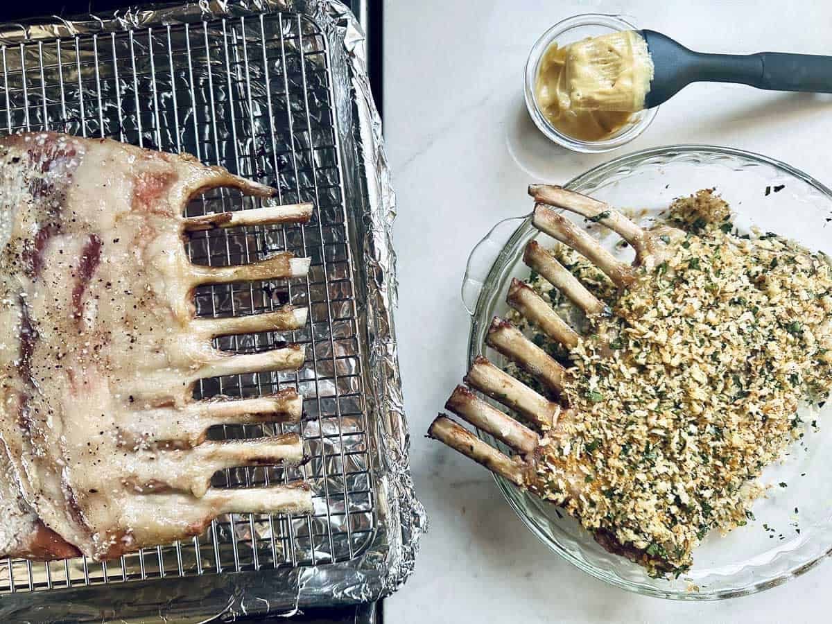 Two racks of lamb: one raw and seasoned on a foil-lined rack, the other as a Crumb-Crusted Rack of Lamb Persillade in a glass dish. A small bowl of mustard with a brush sits nearby on a marble surface.