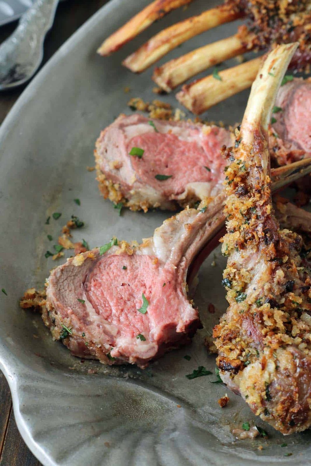 Three slices of Crumb-Crusted Rack of Lamb Persillade, cooked medium-rare, are arranged on a gray serving platter. The lamb is garnished with chopped herbs and has a golden, crispy breadcrumb coating.
