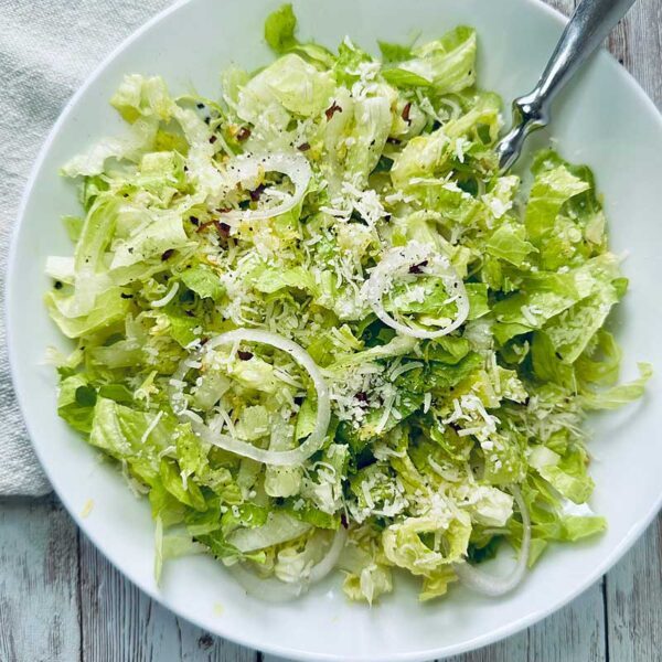 A white bowl filled with Crunchy Romaine Salad—chopped romaine lettuce, thinly sliced onions, grated parmesan cheese, and seasoning—placed on a rustic wooden table with a white napkin and a fork.