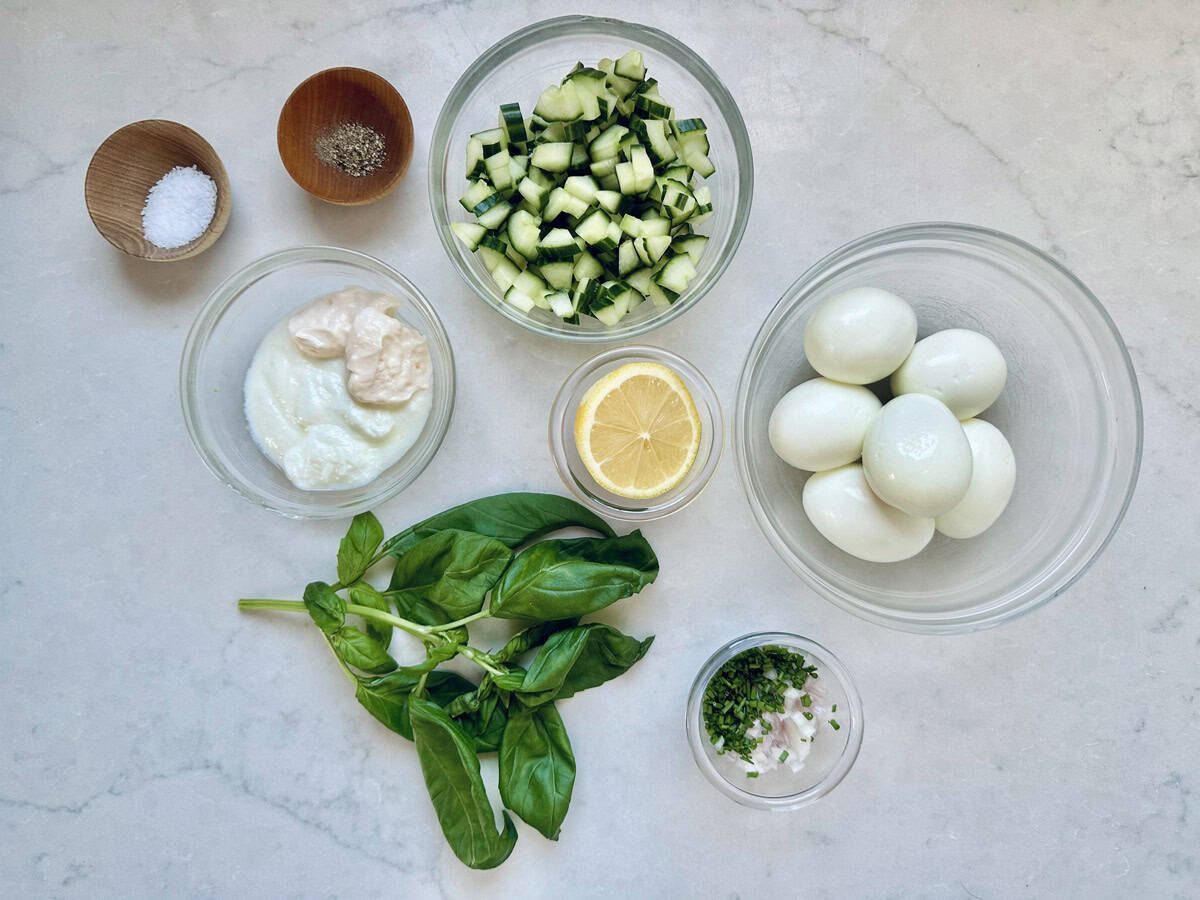 A marble countertop displays bowls of diced cucumber, hard-boiled eggs, mayonnaise and yogurt, chopped herbs with onions, salt and pepper, half a lemon, and fresh basil leaves—ingredients for a refreshing Cucumber and Basil Egg Salad arranged neatly.