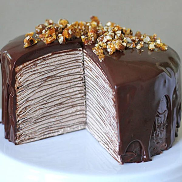 A tall chocolate crepe cake with many thin layers, covered in glossy chocolate ganache and topped with crushed praline, sits on a white cake stand with a large slice removed to show the layered interior.