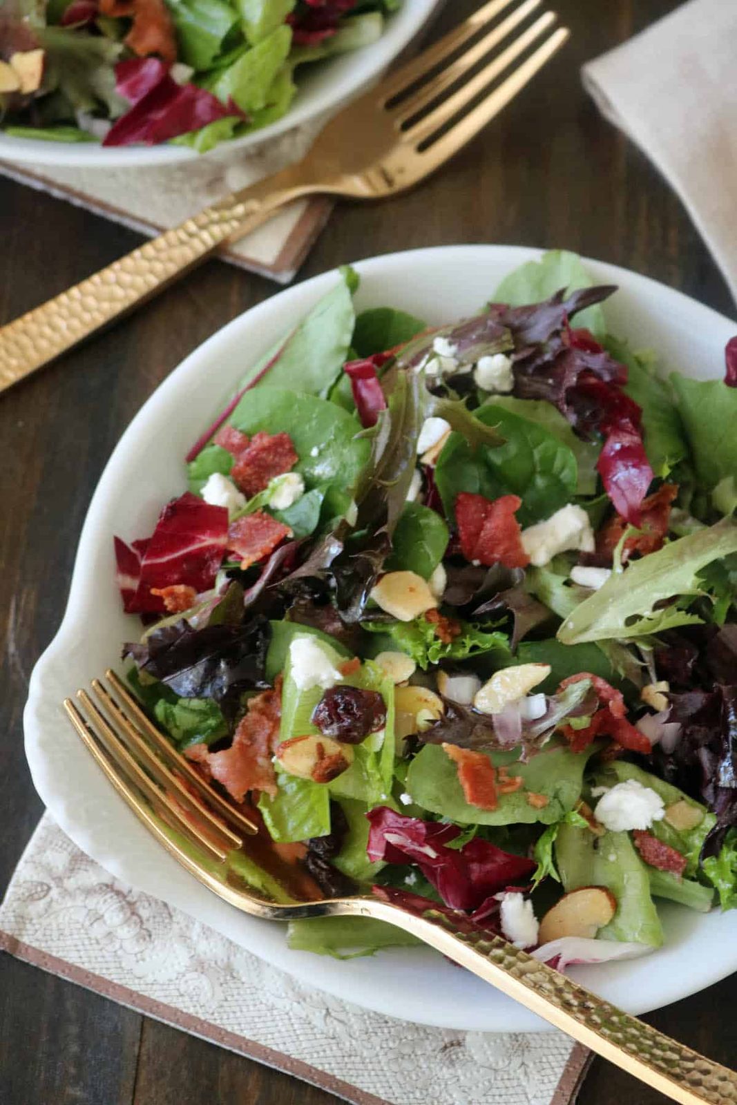 A fresh salad with mixed greens, goat cheese, bacon bits, sliced almonds, and dried cranberries is served in a white bowl with a gold fork on a brown napkin.