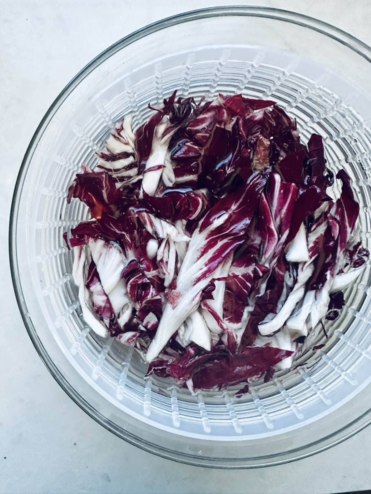 Shredded radicchio leaves in a clear salad spinner, viewed from above, showing their deep purple-red and white colors against a light background.