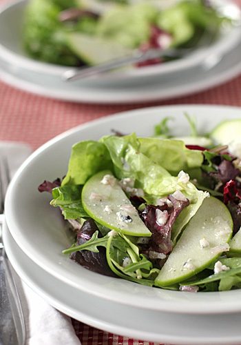 A fresh green salad with mixed leafy greens, sliced green apples, and crumbled cheese served in a white bowl, with another bowl and cutlery visible in the background.