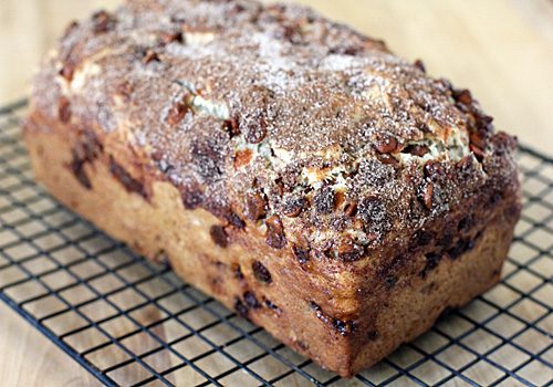 A freshly baked cinnamon nut bread loaf with a golden-brown crust sits on a cooling rack, topped with a sprinkle of cinnamon sugar and visible chunks of nuts.