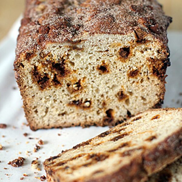 A loaf of cinnamon swirl bread with a sugary, cinnamon-topped crust, partially sliced, resting on parchment paper with crumbs scattered around.