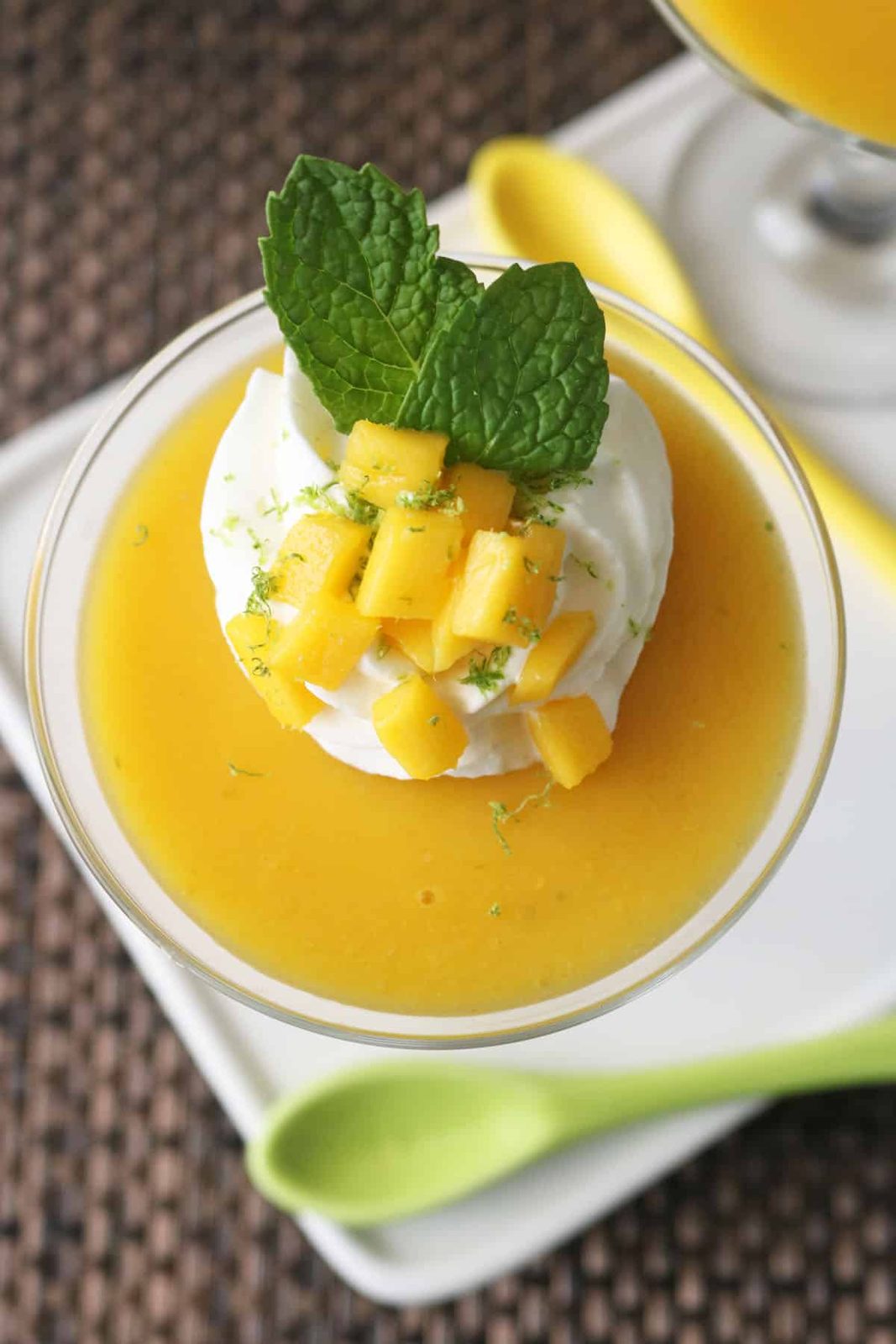 A glass dessert dish filled with smooth mango puree, topped with a swirl of whipped cream, diced mango pieces, lime zest, and two fresh mint leaves, with a green spoon beside the dish.