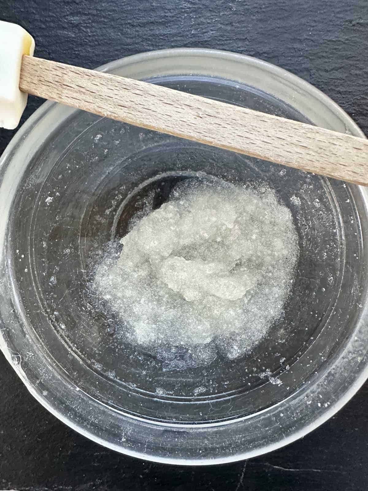 A glass bowl containing a clear, gel-like mixture is shown from above. A wooden spatula with a white silicone tip rests on the rim of the bowl. The background is a dark, textured surface.
