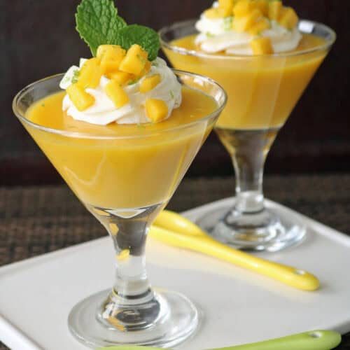 Two glass cups of mango mousse topped with whipped cream, diced mango, and mint leaves, served on a white tray with yellow and green spoons. The background is dark and the setting is elegant.