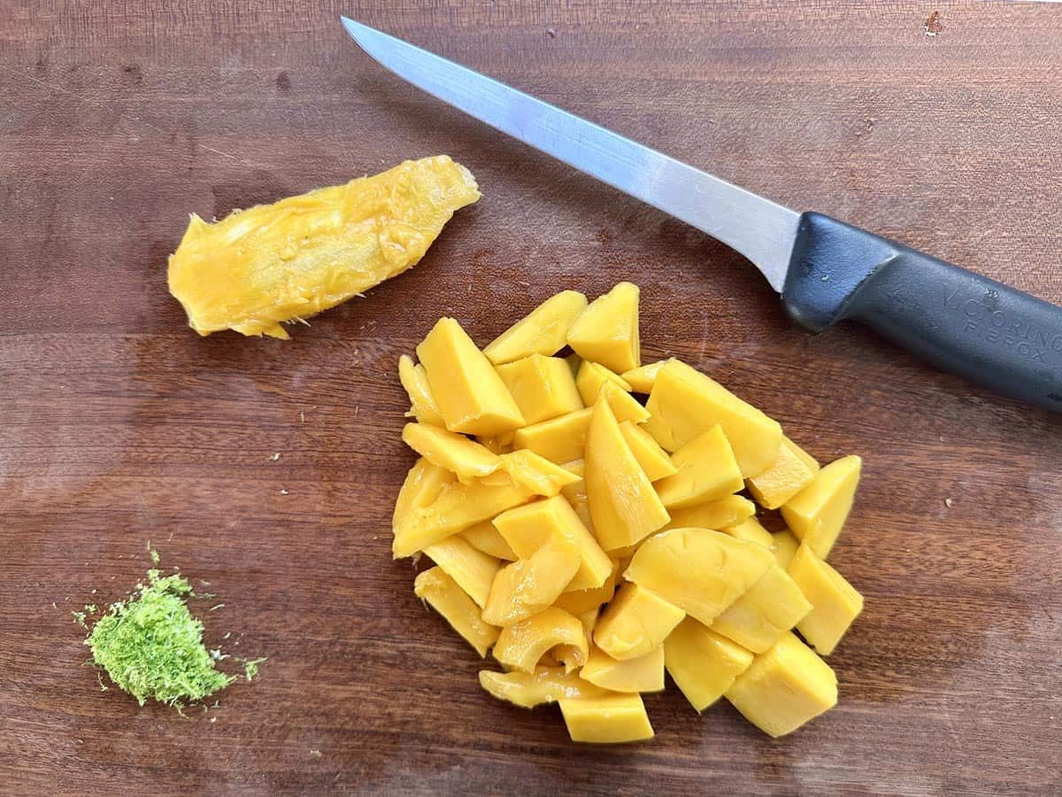A pile of diced mango, a small heap of green lime zest, a mango pit, and a knife with a black handle on a wooden cutting board.