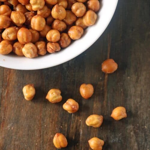 A white bowl filled with roasted chickpeas sits on a dark wooden surface, showcasing this crunchy roasted chickpeas recipe, with several chickpeas scattered outside the bowl.