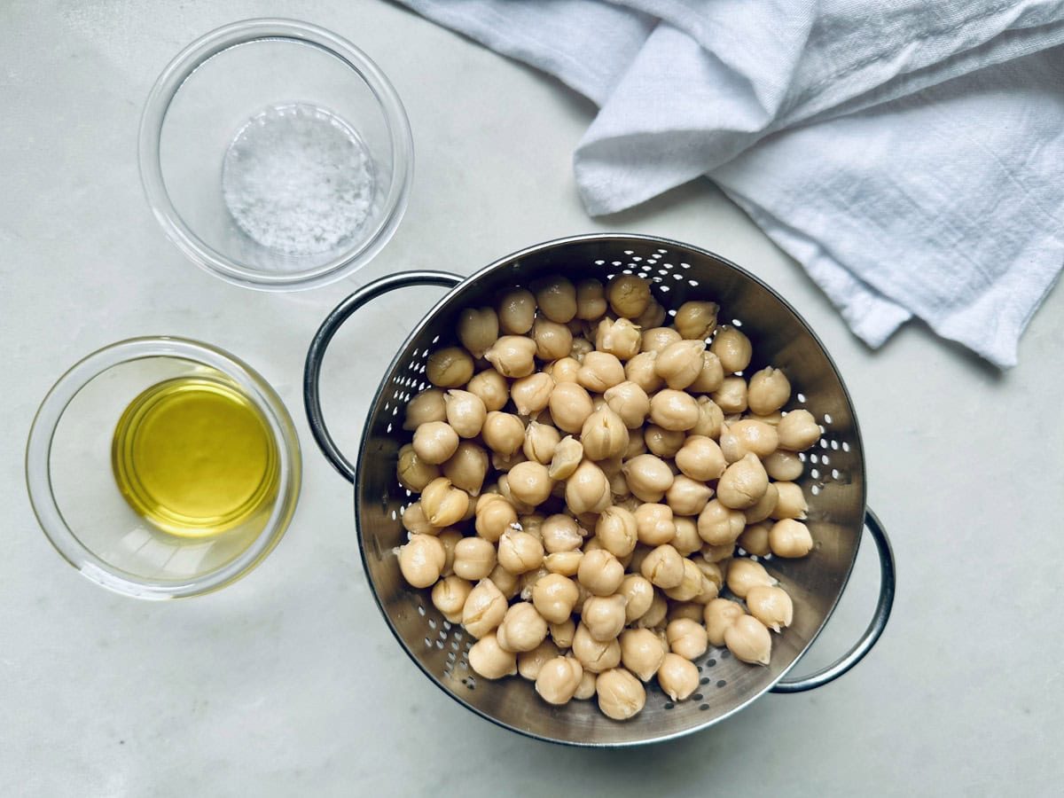 A metal colander filled with chickpeas sits on a light surface beside a small glass bowl of olive oil and a bowl of salt, ready for your roasted chickpeas recipe, with a white cloth in the corner.