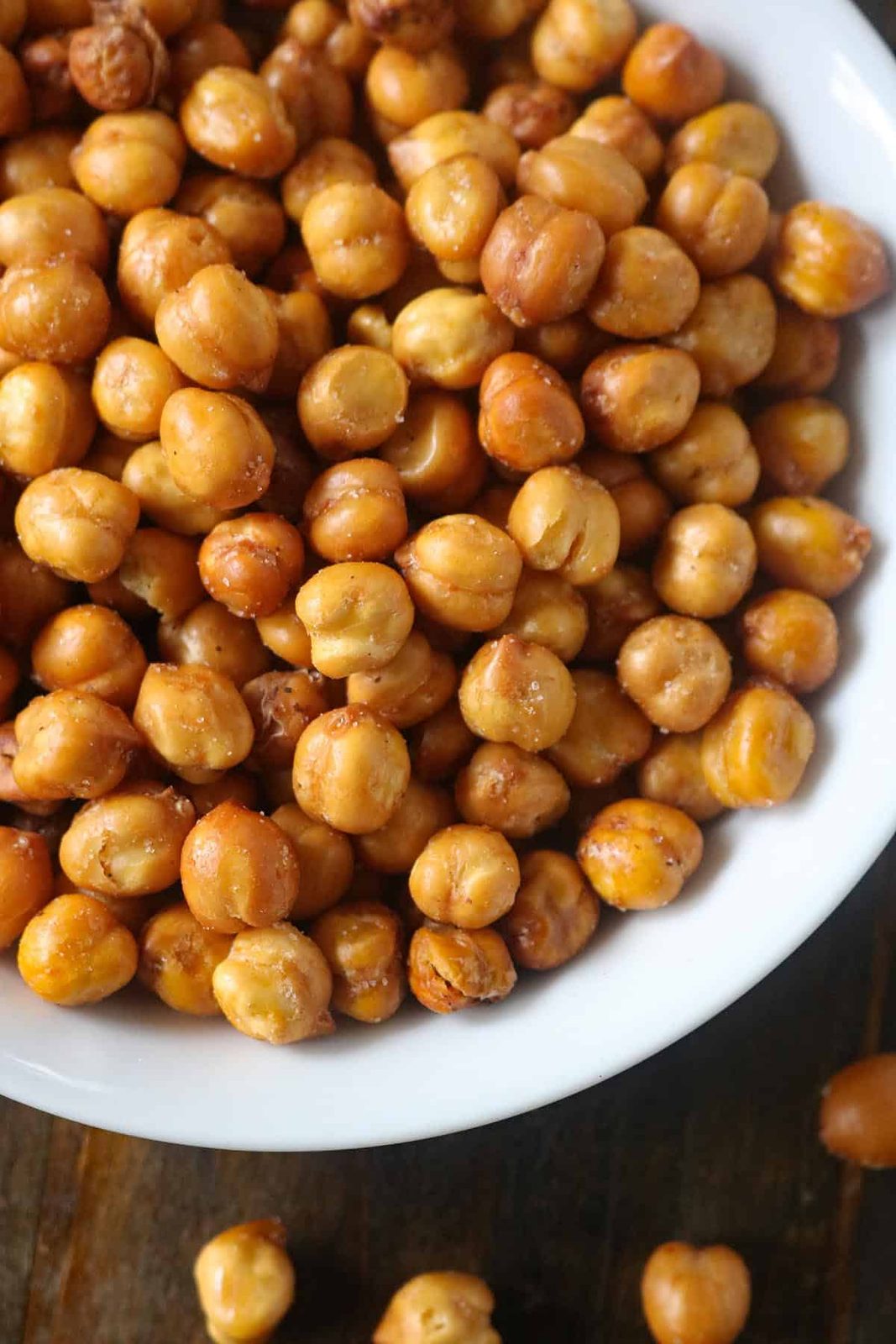 A close-up of a white bowl filled with roasted chickpeas, some golden brown and crispy, sits on a wooden surface with a few scattered around—perfect for showcasing your favorite roasted chickpeas recipe.