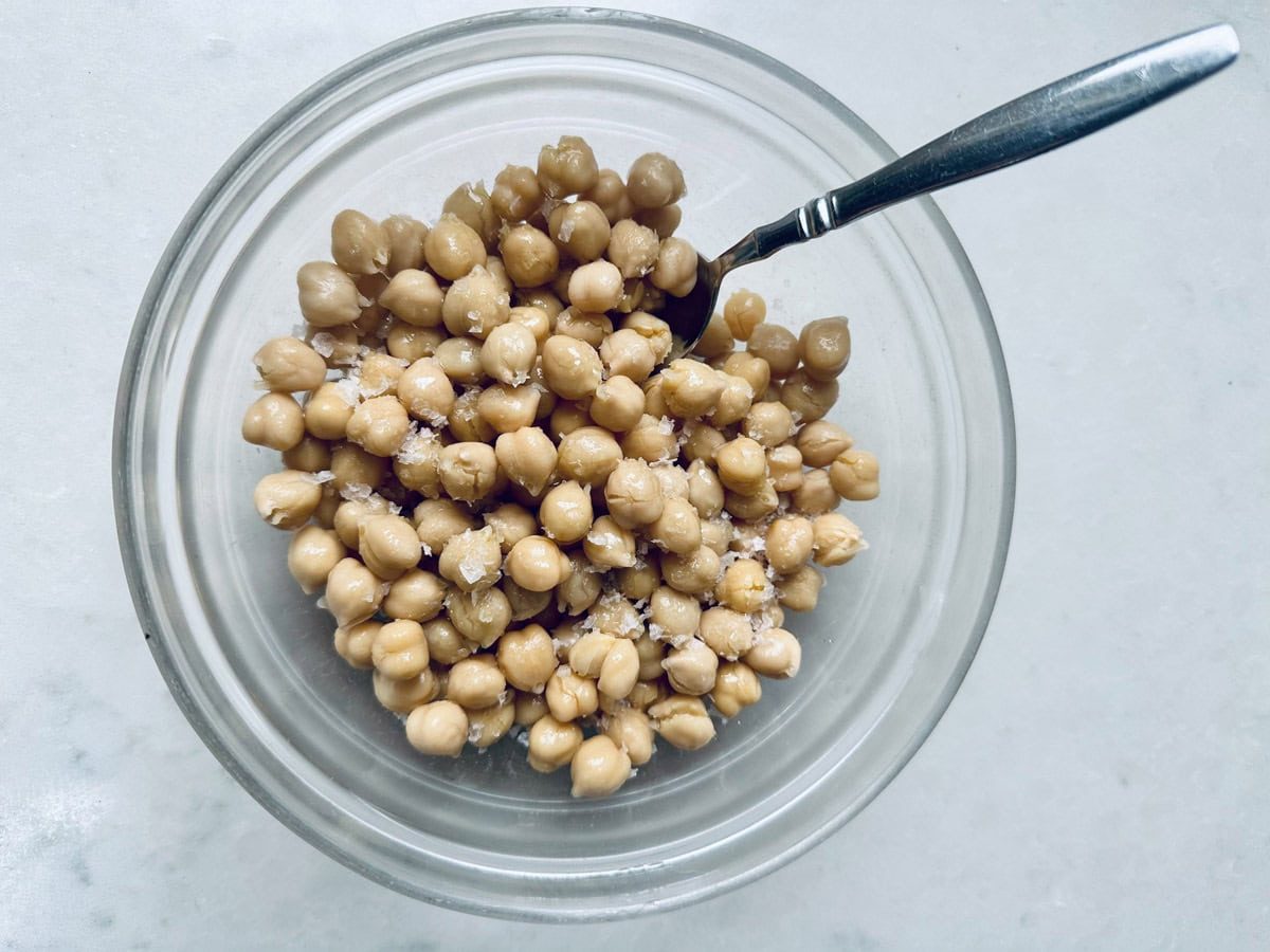 A glass bowl filled with roasted chickpeas topped with coarse salt, with a metal spoon resting inside, placed on a light-colored surface—a simple and savory roasted chickpeas recipe.