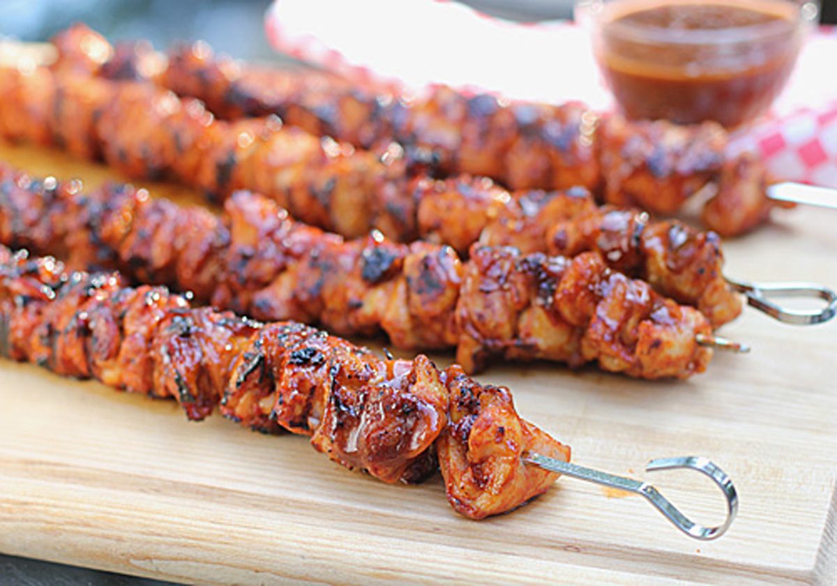 Four grilled meat skewers, coated in a shiny, reddish sauce, rest on a wooden cutting board. A small bowl of dipping sauce is visible in the background.