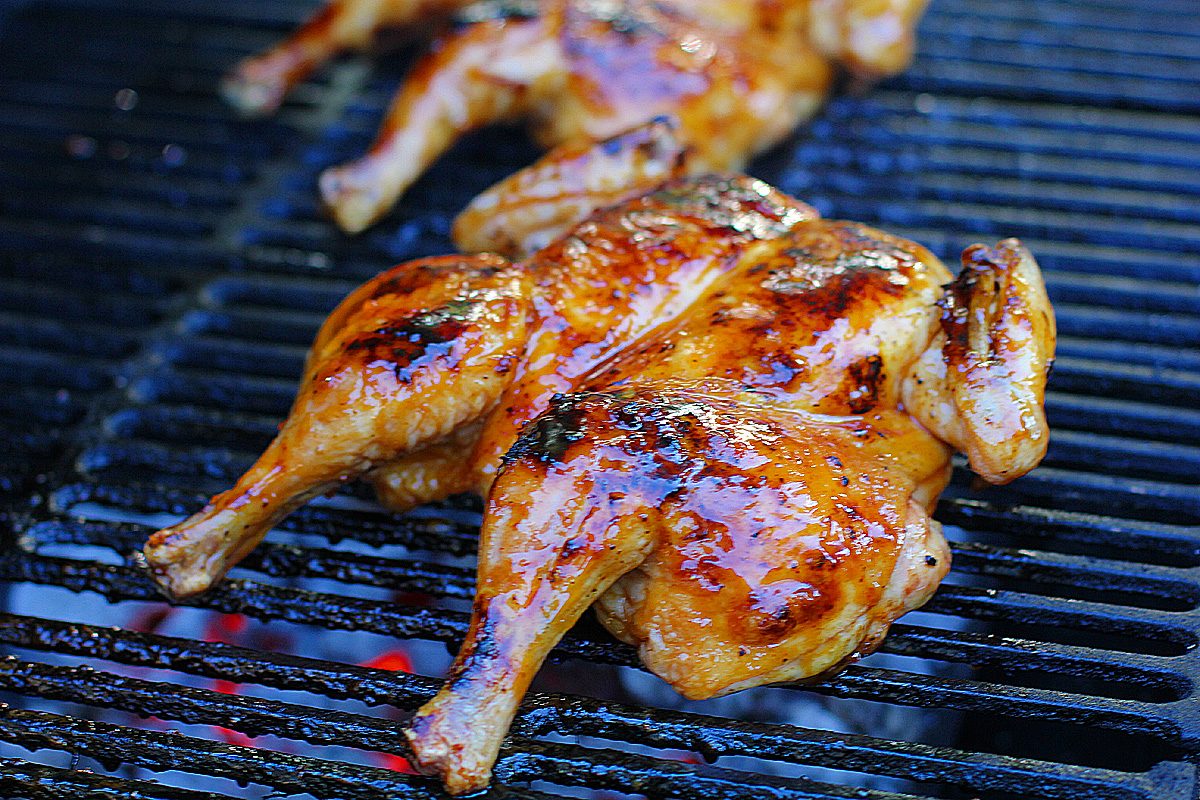 Grilled, glazed chickens are cooking on a barbecue grill, showing golden brown, slightly charred skin with a shiny, caramelized coating.