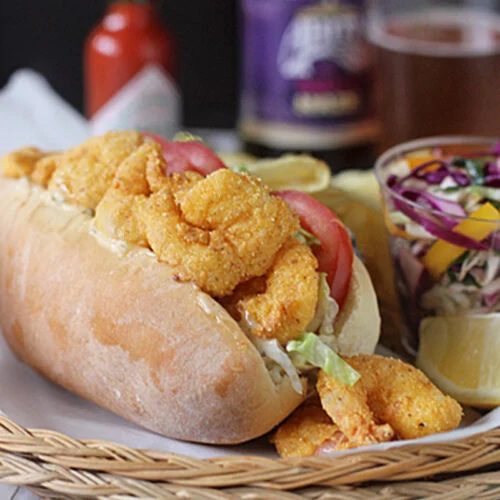 A shrimp po’ boy sandwich with lettuce and tomato on a hoagie roll sits on a plate with coleslaw, lemon wedges, and potato chips. Mardi Gras beads, hot sauce, beer, and soft drink bottles are in the background.