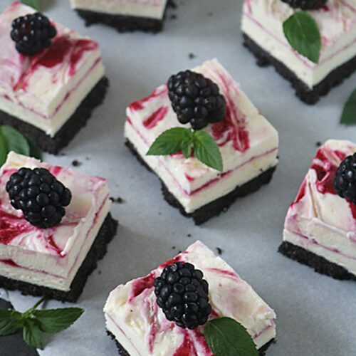 Square cheesecake bars with a chocolate cookie base, swirled berry sauce on top, each garnished with a fresh blackberry and green mint leaves, arranged on parchment paper.