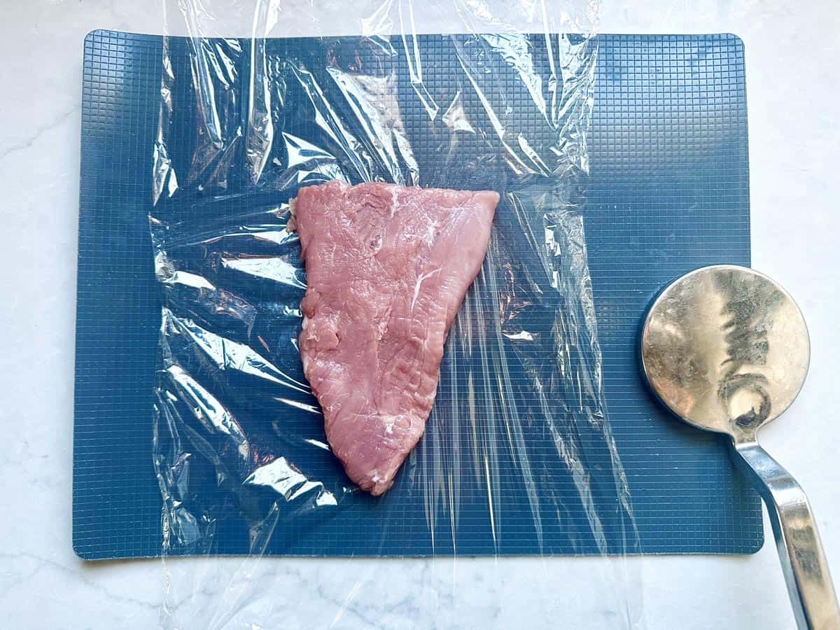 A raw piece of meat sits on a blue cutting board, partially covered with plastic wrap. A metal meat tenderizer is placed to the right of the meat.