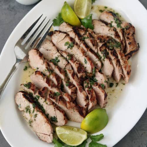 Sliced grilled pork garnished with chopped herbs and served with lime wedges and cilantro on a white plate, with a fork and a small jug of sauce on the side.