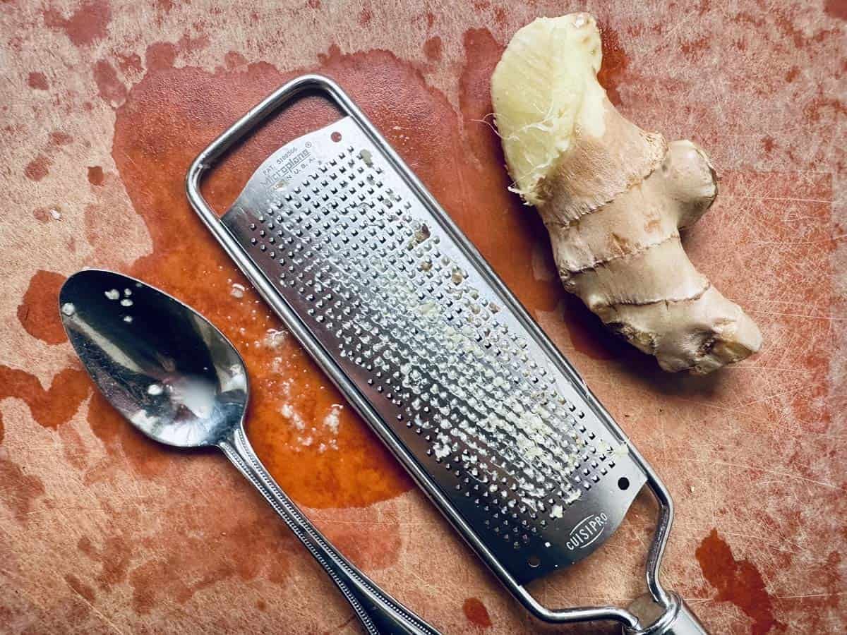 A metal grater, a spoon with some grated ginger, and a piece of fresh ginger root are on a reddish cutting board with juice stains.