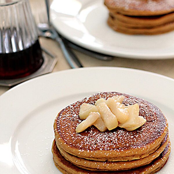 A stack of pancakes topped with diced fruit and powdered sugar sits on a white plate. In the background, another plate of pancakes and a glass syrup dispenser are visible.
