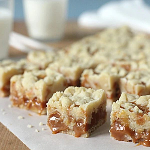 Close-up of caramel crumb bars cut into squares on parchment paper, with two glasses of milk in the background on a wooden surface.