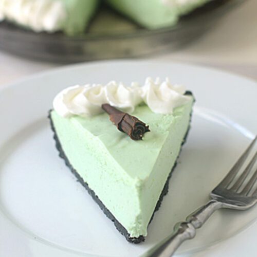 A slice of light green pie with a chocolate cookie crust, topped with whipped cream and a chocolate curl, sits on a white plate next to a fork. The rest of the pie is in the background.