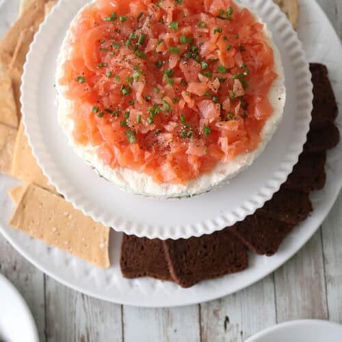A plate with a round Gravlax Torta—smoked salmon and cream cheese dip topped with chives—surrounded by assorted crackers and bread slices on a rustic wooden table.