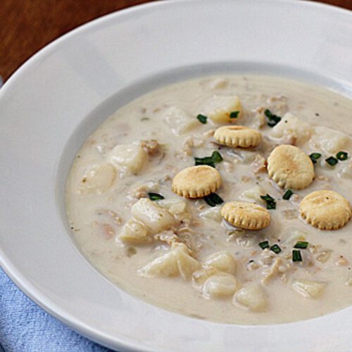A bowl of creamy clam chowder with chunks of potatoes, garnished with chopped chives and round oyster crackers, served on a white plate next to a spoon on a blue napkin.