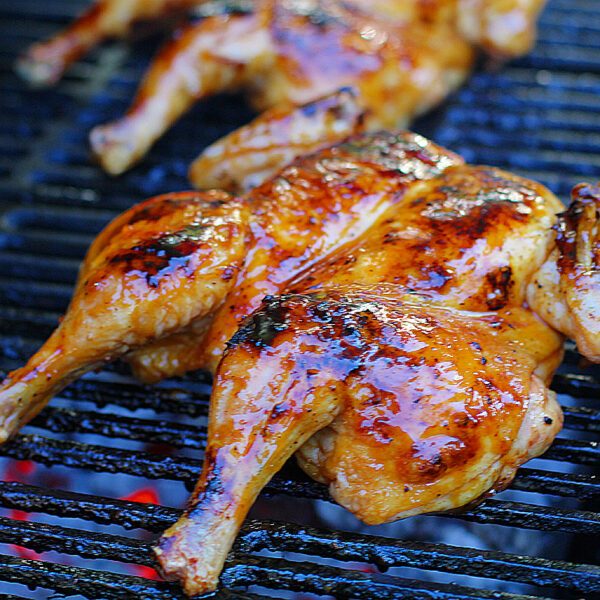 Grilled, glazed chickens are cooking on a barbecue grill, showing golden brown, slightly charred skin with a shiny, caramelized coating.