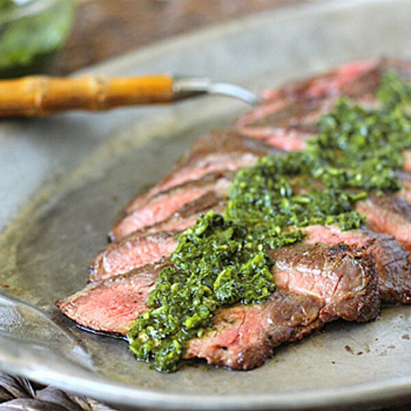 Sliced medium-rare steak arranged on a gray plate, topped with a green chimichurri sauce, with a serving utensil and a bowl of extra sauce in the background.