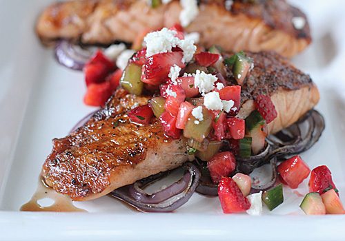 Grilled salmon fillet served on a bed of red onion slices, topped with a colorful strawberry salsa and crumbled feta cheese on a white plate.