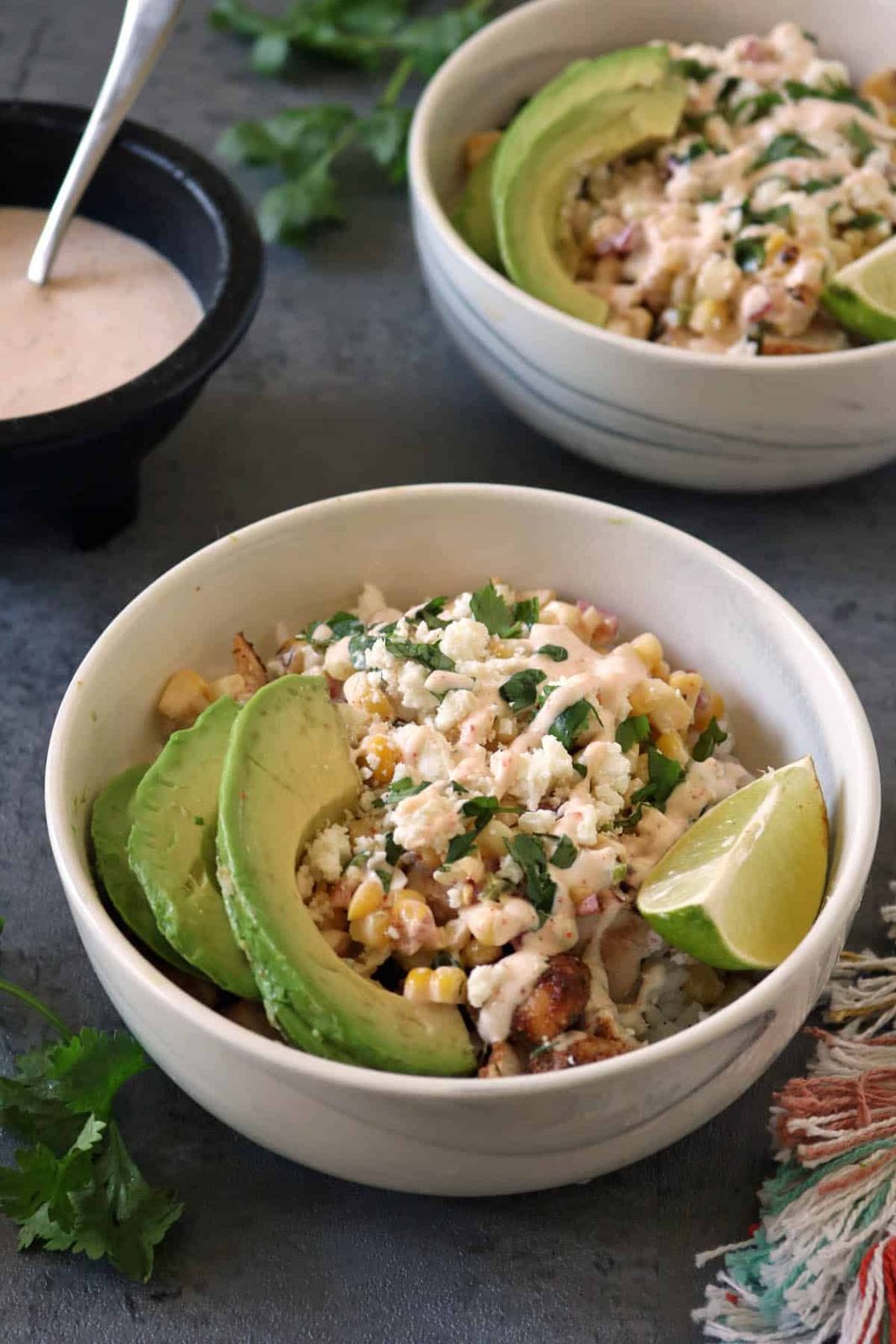 A bowl of grilled corn salad topped with avocado slices, crumbled cheese, chopped herbs, and a lime wedge, with a drizzle of creamy dressing—just like the flavors in classic Grilled Street Corn Chicken Bowls. Another bowl and dressing bowl are in the background.