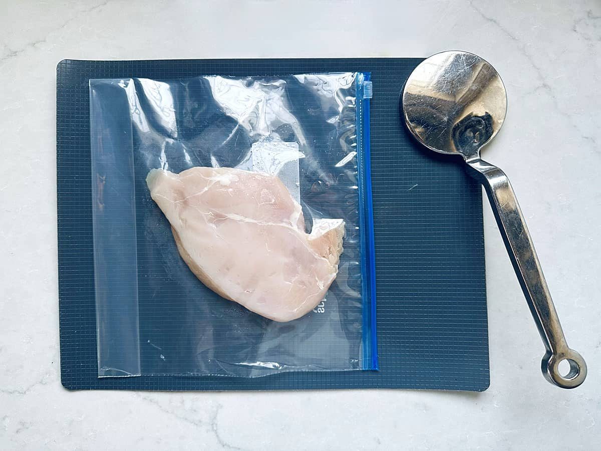 A raw chicken breast in a resealable plastic bag on a dark cutting board, next to a metal kitchen mallet on a white countertop—ready to be tenderized for Grilled Street Corn Chicken Bowls.