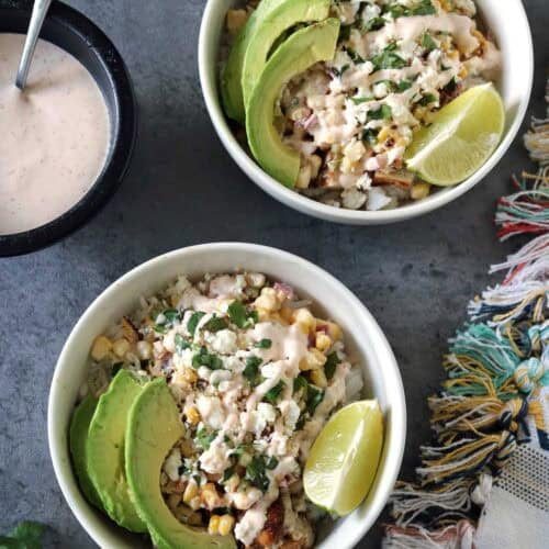 Two bowls of Grilled Street Corn Chicken Bowls topped with avocado slices, lime wedges, cheese, and creamy dressing sit on a gray surface, accompanied by a colorful cloth, fresh cilantro, and extra dressing on the side.