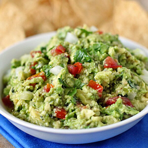 A white bowl filled with chunky guacamole, containing diced tomatoes, onions, and cilantro, sits on a blue cloth with tortilla chips visible in the background.