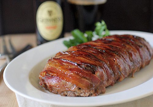 A bacon-wrapped meatloaf sits on a white plate, garnished with fresh herbs, with a bottle of stout beer and utensils blurred in the background.