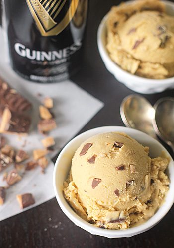 Two bowls of ice cream with chocolate chunks, next to two spoons, pieces of toffee candy, and a can of Guinness beer on a dark surface.